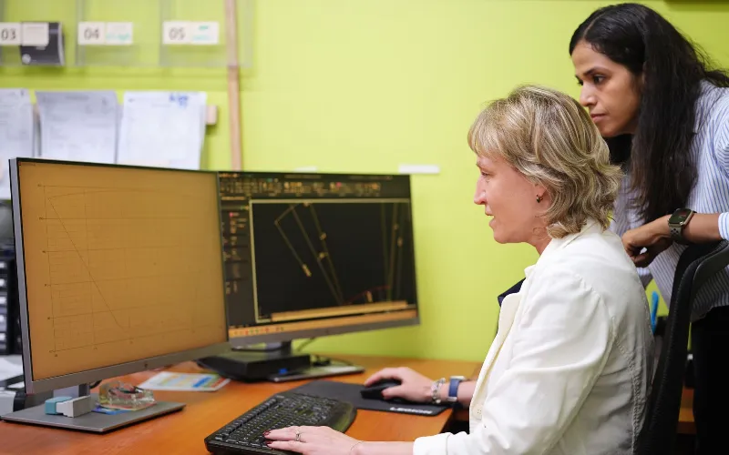 Two professionals reviewing technical data and CAD diagrams on dual computer monitors in an engineering office.