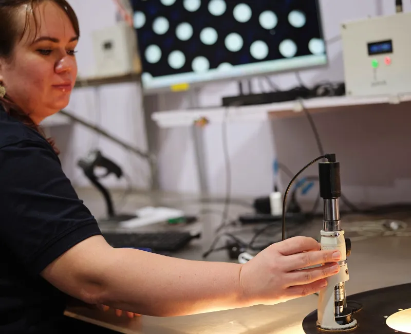 Technician adjusting an optical measurement device to inspect a circular metal filtration component under controlled lighting.